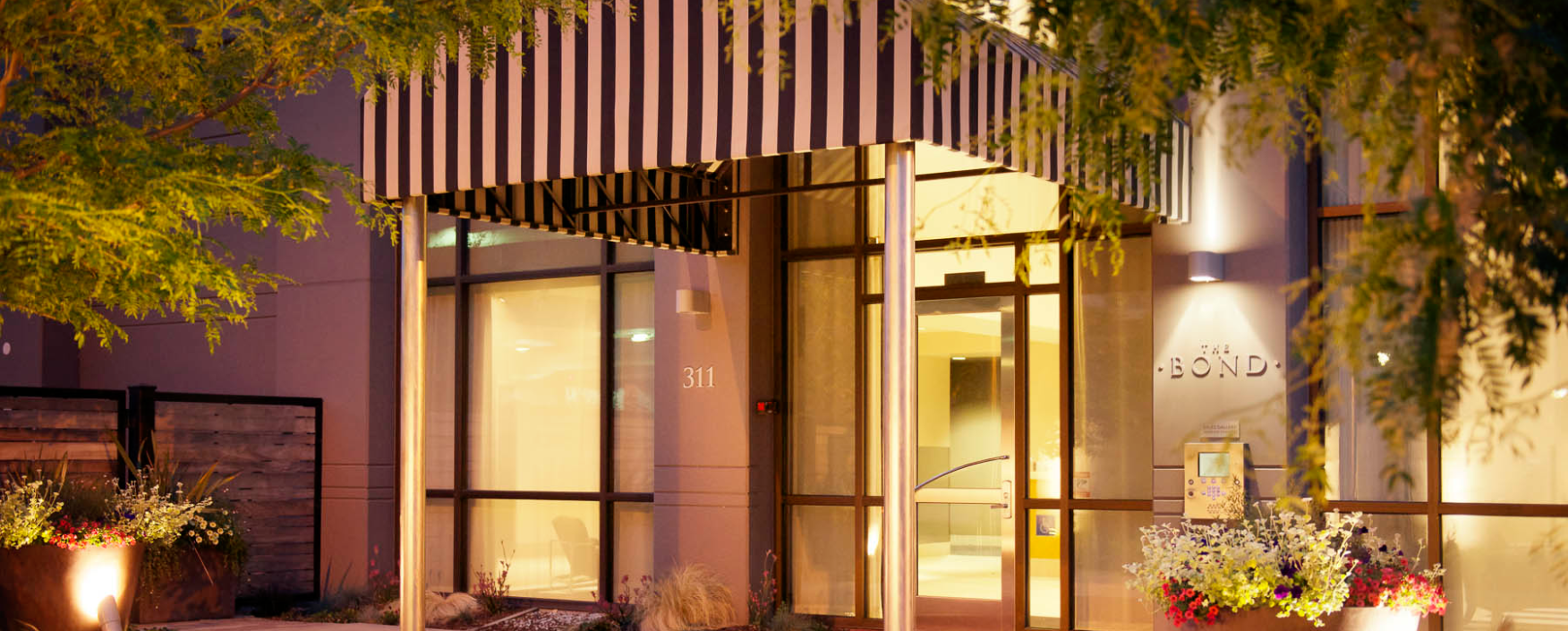 Entrance to a building with a black and white striped awning, glass doors, windows, lights, a name board, plants
