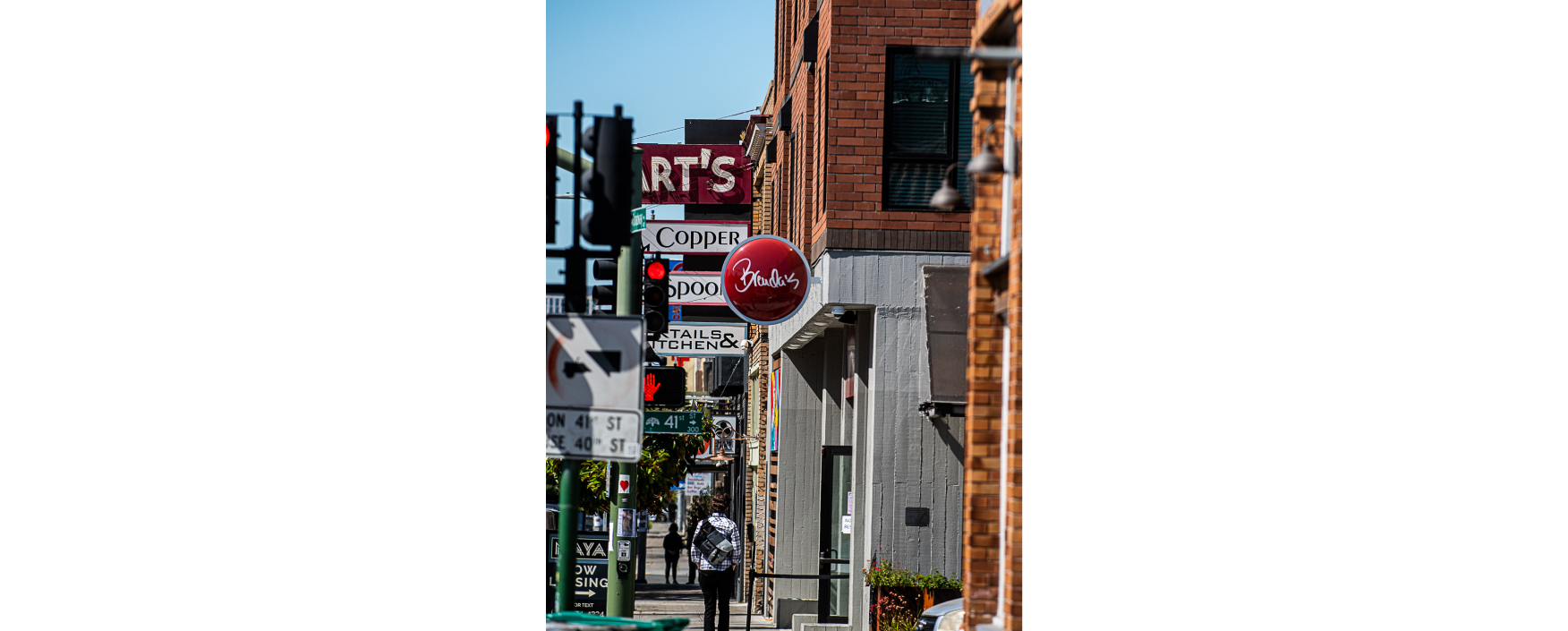 Person walking down a street lined with tall buildings, with a traffic light and sign board in the foreground