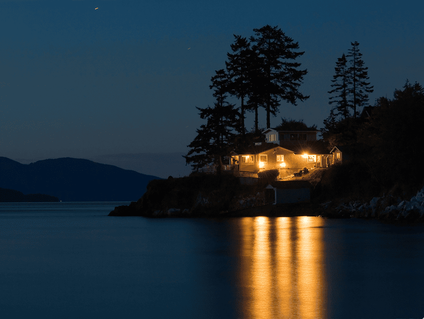 House on the shore of a body of water at night, illuminated by lights and surrounded by trees